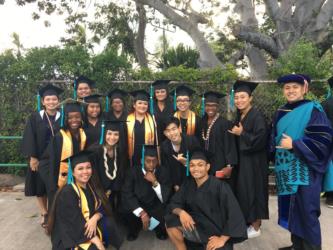 graduates in caps and gowns posing outdoors with a faculty member