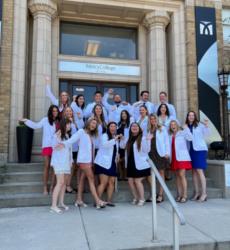 group of students in white coats on steps, smiling and waving